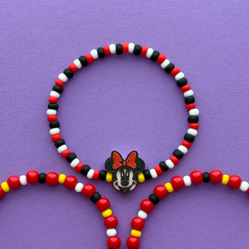 May include: Three beaded bracelets with a Minnie Mouse theme. The bracelets feature red, black, white, and yellow beads. One bracelet has a Minnie Mouse charm. The bracelets are on a purple background.