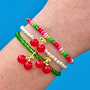 May include: A set of four bracelets with red cherry charms. The bracelets are made of green, pink, and white beads.