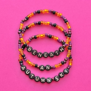 May include: Three beaded bracelets with black letter beads spelling out "AUTUMN SKY", "AN", and "BENEATH". The beads are orange, black, and purple.