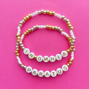 May include: Two beaded bracelets with gold and silver beads. The bracelets spell out the words "INOMNIA PARATUS" in gold letters on white beads.