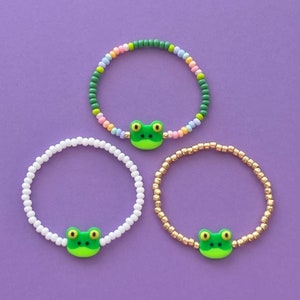 May include: Three beaded bracelets with frog charms. The bracelets are white, gold, and multi-colored.