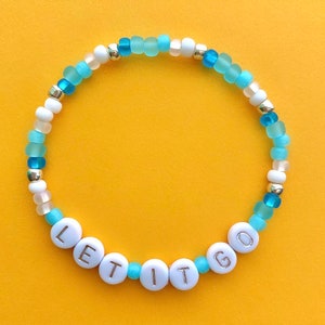 Könnte beinhalten: Ein Perlenarmband mit den Worten "LET IT GO" in weißen Buchstabenperlen. Das Armband hat eine Mischung aus blauen, weißen und goldfarbenen Perlen, vor einem gelben Hintergrund.