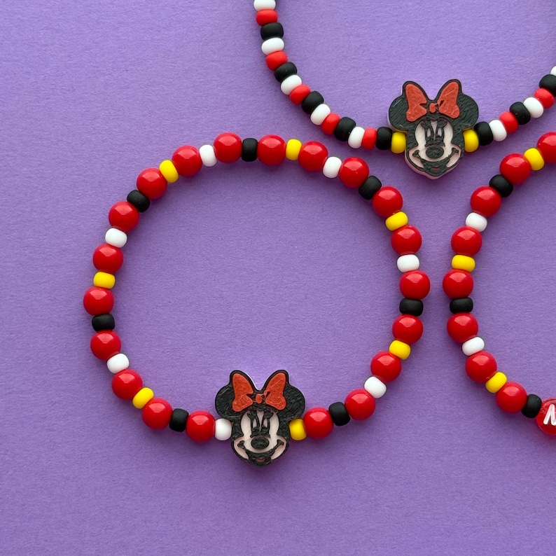 May include: Three beaded bracelets with a Minnie Mouse charm. The bracelets feature red, black, white, and yellow beads. The Minnie Mouse charm is black with a red bow and yellow accents. The bracelets are on a purple background.