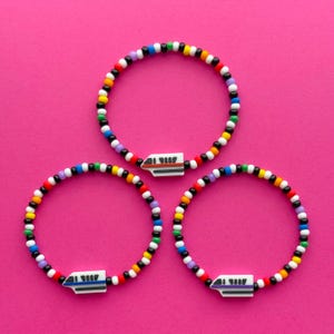 Könnte beinhalten: Drei Armbänder mit Perlen mit einem farbenfrohen Muster aus schwarzen, weißen, roten, gelben, grünen, lila und blauen Perlen. Jedes Armband hat einen weißen Anhänger mit einem Zugdesign. Die Armbänder werden auf einem rosa Hintergrund präsentiert.