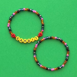 May include: Two beaded bracelets with a colorful mix of black, brown, pink, and blue beads. One bracelet has the word "GAMBIT" spelled out in yellow beads with red "X" beads on either side.