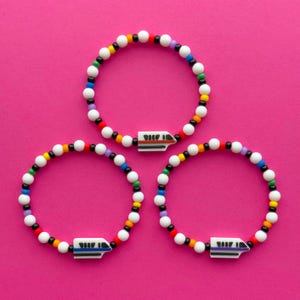 Könnte beinhalten: Drei Armbänder mit Perlen in Weiß, Schwarz und Regenbogenfarben. Jedes Armband hat einen weißen Zuganhänger mit roten, schwarzen und blauen Details. Die Armbänder werden auf einem hellrosa Hintergrund präsentiert.