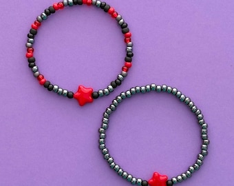 Puede incluir: Dos pulseras de cuentas. Una pulsera presenta cuentas rojas, negras y grises con un dije de estrella roja. La otra pulsera tiene cuentas grises y un dije de estrella roja. Ambas pulseras están sobre un fondo morado.
