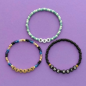 May include: Three beaded bracelets on a purple background. One bracelet is teal with the name "BOB" spelled out in white letter beads. Another bracelet is black with "THE VOID" spelled out in white letter beads. The third bracelet is blue, gold, and black with "SENTRY" spelled out.