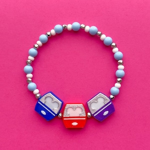 Könnte beinhalten: Ein Armband mit hellblauen, weißen und silbernen Perlen. Das Armband hat drei quadratische Anhänger in Blau, Rot und Lila, jeder mit einem silbernen Herzdesign. Das Armband befindet sich auf einem rosa Hintergrund.
