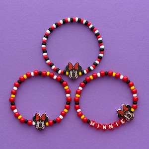May include: Three beaded bracelets with a Minnie Mouse theme. The bracelets feature red, black, white, and yellow beads. One bracelet spells "Minnie" with white beads. Each bracelet has a Minnie Mouse charm.