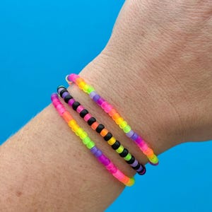 May include: Three beaded bracelets in vibrant neon colors are displayed on a wrist. The bracelets feature a variety of colors, including pink, orange, yellow, green, purple, and black. The beads are small and evenly spaced.