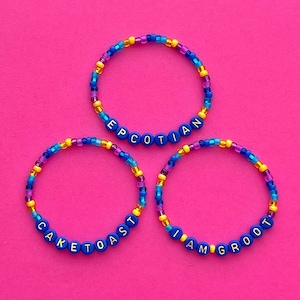 May include: Three beaded bracelets with different words spelled out on them. The bracelets are blue and yellow with purple beads. The words on the bracelets are "Epcotian", "Cake Toast", and "I Am Groot".