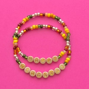 Peut inclure: Deux bracelets en perles avec les mots "HAPPY" et "WINDY DAY" écrits en lettres dorées sur des perles beiges. Les bracelets sont faits de petites perles colorées dans les tons jaunes, orange, rouge, vert et argent.