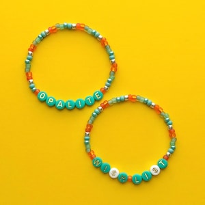 Puede incluir: Dos pulseras de cuentas con las palabras "OPALITE" y "WISH LITE" escritas con cuentas de letras turquesas. Las pulseras tienen cuentas naranjas, plateadas y turquesas. El fondo es amarillo liso.