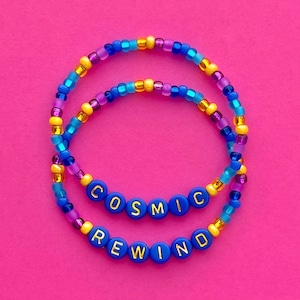 May include: Two beaded bracelets with blue, purple, and gold beads. The bracelets spell out "COSMIC" and "REWIND" in blue letters.