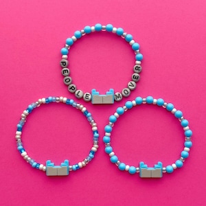 Könnte beinhalten: Drei Armbänder mit Perlen auf pinkfarbenem Hintergrund. Die Armbänder haben blaue und weiße Perlen. Eines der Armbänder schreibt "PEOPLE MOVER" mit Buchstabenperlen. Jedes Armband hat einen hellblau-grauen Anhänger.