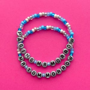 Könnte beinhalten: Zwei Armbänder mit Perlen mit den Worten "PAGING MR TOMORROW" in silbernen Buchstabenperlen. Die Armbänder haben eine Mischung aus blauen, weißen und silbernen Perlen. Der Hintergrund ist ein helles Pink.