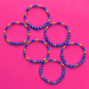 May include: Six colorful beaded bracelets with different words spelled out in gold letters on blue beads. The words are "CONGA", "SEPTEMBER", "DISCO", "INFERNO", "I RAN ONE WAY OR ANOTHER", and "RULE THE WORLD".