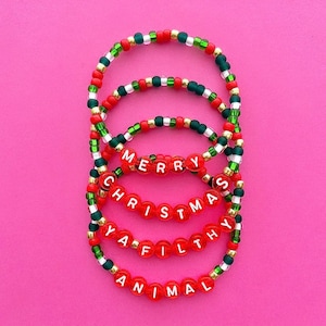 May include: Four red beaded bracelets with white lettering spelling out "Merry Christmas Ya Filthy Animal". The bracelets are layered on top of each other and have green and gold beads.