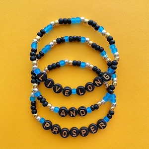 Puede incluir: Tres pulseras de cuentas con cuentas negras y doradas y detalles azules. Las pulseras deletrean "Live Long and Prosper".