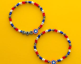 Puede incluir: Dos pulseras de cuentas rojas, blancas, azules y grises. Una pulsera deletrea "CAP" con cuentas de letras y presenta un dije de escudo del Capitán América. La otra pulsera tiene un dije de escudo del Capitán América. Las pulseras están sobre un fondo amarillo.