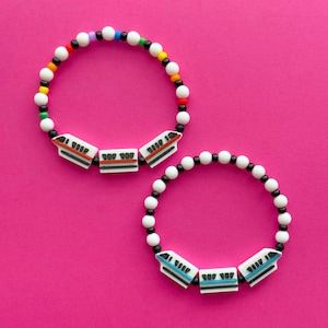 Könnte beinhalten: Zwei Armbänder mit Perlen auf pinkfarbenem Hintergrund. Jedes Armband hat weiße, schwarze und farbige Perlen. Die Armbänder haben weiße, rechteckige Anhänger mit einem Zugdesign. Eines hat orangefarbene Akzente, das andere blaue Akzente.