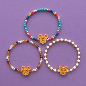 May include: Three beaded bracelets with a Mickey Mouse charm on a purple background. One bracelet features colorful beads, another has brown and gold beads, and the third has white and gold beads. The charms are a light brown color.