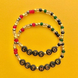 May include: Two beaded bracelets with gold, black, green, and red beads. The bracelets spell out "Let's Do Get The Help".