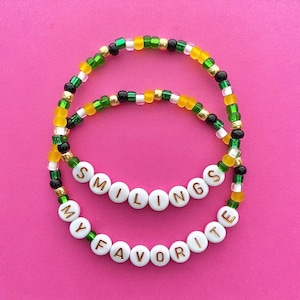 Op de afbeelding: Twee kralenarmbanden met groene, gele en zwarte kralen. De armbanden spellen "MY FAVORITE SMILINGS" in gouden letters op witte kralen.