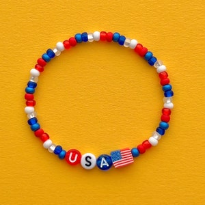 Puede incluir: Un brazalete de cuentas rojo, blanco y azul con las letras "USA" y un pequeño dije de bandera estadounidense.