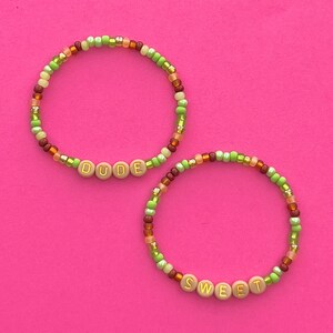 May include: Two beaded bracelets with the words "DUDE" and "SWEET" spelled out in gold letters on brown beads. The bracelets are made with green, orange, and brown beads.