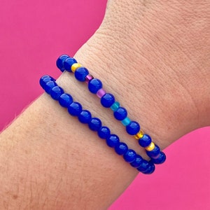 May include: Two beaded bracelets on a wrist. The bottom bracelet is made of solid blue beads. The top bracelet has blue, yellow, pink, and light blue beads. The background is a bright pink.