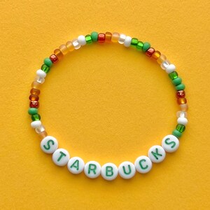 May include: A white beaded bracelet with green and brown beads. The beads spell out the word "STARBUCKS" in green letters.