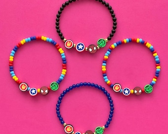 Puede incluir: Tres pulseras de cuentas con dijes de personajes de superhéroes. Las pulseras son negras, azules y rojo, amarillo y azul. Cada pulsera tiene una cuenta dorada, un escudo rojo y azul, una araña roja y un puño verde.