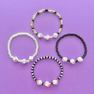 Puede incluir: Cuatro pulseras con dijes de dados. Las pulseras están hechas de cuentas negras, verdes y blancas. Los dijes de dados son blancos con puntos negros.