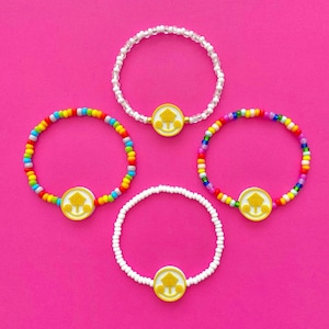 May include: Four colorful beaded bracelets with a yellow charm featuring a stylized tree design. The bracelets are on a pink background.