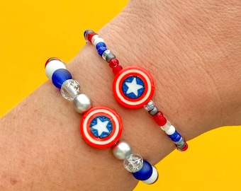 Puede incluir: Dos pulseras de cuentas rojas, blancas y azules. Cada pulsera tiene un dije redondo con una estrella blanca sobre fondo azul, rodeada por un círculo rojo. Las pulseras están sobre un fondo amarillo.