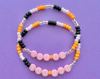 May include: Two beaded bracelets with alternating orange, black, and white beads. One bracelet spells out "YOU DO" in pink beads with orange spacers. The other bracelet spells out "YOU BOO" in pink beads with orange spacers.