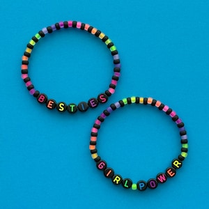 Könnte beinhalten: Zwei Armbänder mit Perlen mit den Wörtern "BESTIES" und "GIRL POWER". Die Armbänder haben einen Regenbogenfarbverlauf und schwarze Perlen. Die Buchstaben sind in leuchtenden Farben gehalten. Der Hintergrund ist einfarbig blau.
