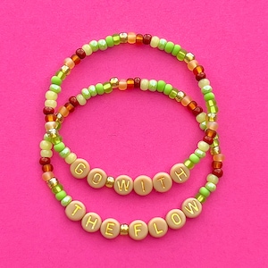 Può includere: Due braccialetti di perline con la frase "Go With The Flow" scritta in lettere dorate su perline beige. I braccialetti sono realizzati con perline verdi, arancioni e rosse.