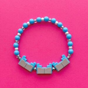 Könnte beinhalten: Ein Armband mit hellblauen und silbernen Perlen, sowie drei hellblauen und grauen Dekorationselementen. Das Armband befindet sich vor einem leuchtend pinkfarbenen Hintergrund, was einen lebendigen Kontrast erzeugt.