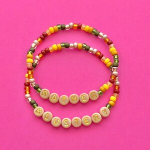 Peut inclure: Deux bracelets en perles avec les mots "Rather" et "Blustery" écrits en lettres dorées sur des perles beiges. Les bracelets sont faits de perles rouges, jaunes, orange, vertes et argentées.
