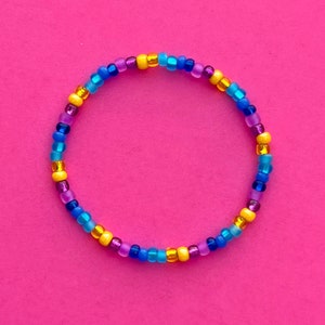 May include: A colorful beaded bracelet with blue, purple, and gold beads. The bracelet is on a pink background.