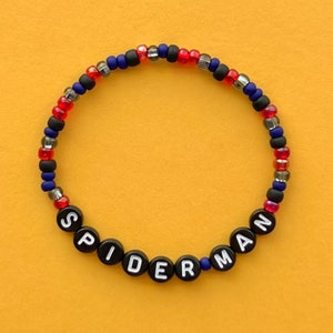 Può includere: Un braccialetto di perline con perline nere, rosse e blu che compongono la scritta "SPIDERMAN".