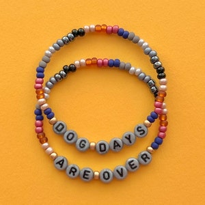 May include: Two beaded bracelets with the words "Dog Days Are Over" spelled out in black letters on gray beads. The bracelets are made with a variety of colors, including gray, black, blue, red, and orange.