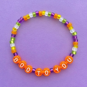 Puede incluir: Un brazalete de cuentas de colores con las palabras "BOO TO YOU" escritas en letras blancas sobre cuentas naranjas. El brazalete está hecho con cuentas moradas, verdes, blancas y naranjas.