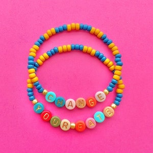 Puede incluir: Dos pulseras de cuentas con las palabras "Change Your Fate" escritas en cuentas de colores. Las pulseras están hechas de cuentas azules y amarillas.