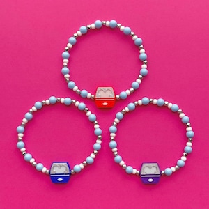 Könnte beinhalten: Drei elastische Armbänder mit blauen, weißen und silbernen Perlen. Jedes Armband hat einen Charm in Form einer Gondel: rot, blau und lila. Die Armbänder werden auf einem hellrosa Hintergrund präsentiert.