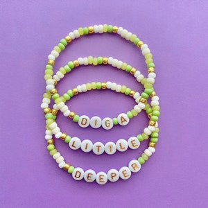 May include: Three beaded bracelets with the words "DIGA", "LITTLE", and "DEEPER" spelled out in gold letters on white beads. The bracelets are made with white and light green beads.