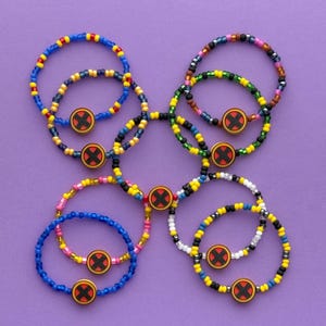 Puede incluir: Una colección de pulseras de cuentas en varios colores, incluyendo azul, amarillo, rosa y negro. Cada pulsera presenta un dije circular con una X roja sobre un fondo amarillo. Las pulseras están dispuestas sobre una superficie morada.